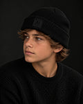 Young person wearing the Possibility Beanie in the Small Fit, black, chunky rib knit with the embroidered Ai element tile on the cuff, dark studio