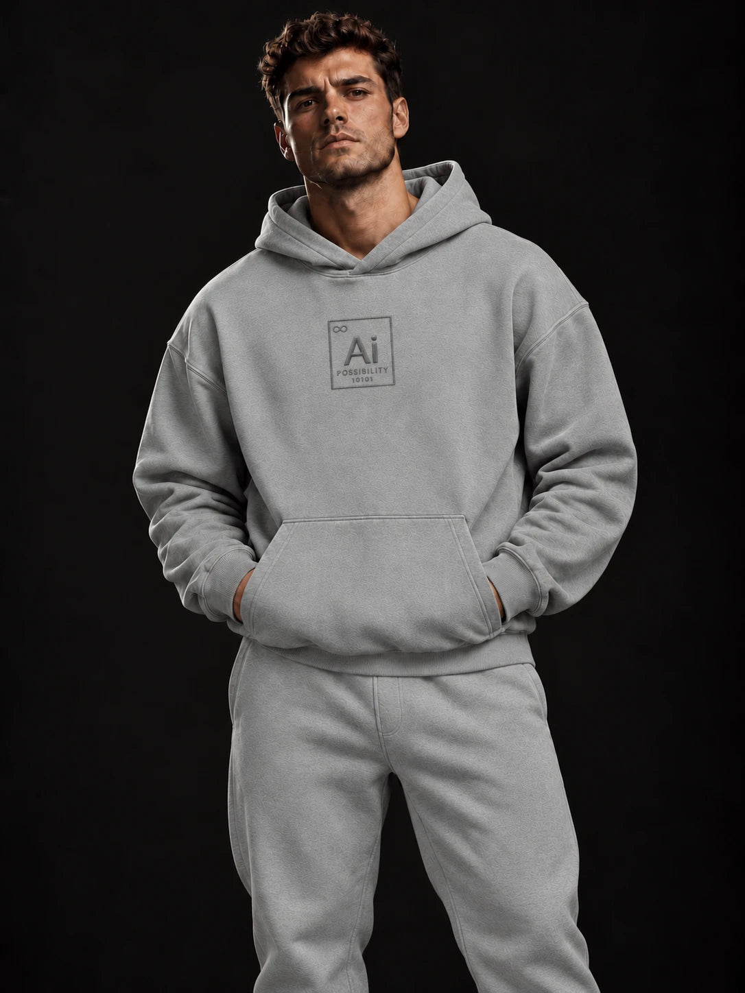 Model wearing the Possibility Hoodie in misty grey with matching joggers, grey-on-grey embroidered Ai element tile on the chest, dark studio